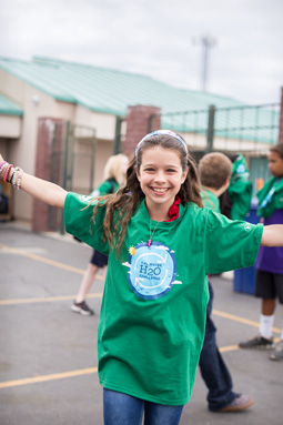 Photograph of a 5th grade girl, arms outstretched, smiling.  A group of children in the background.